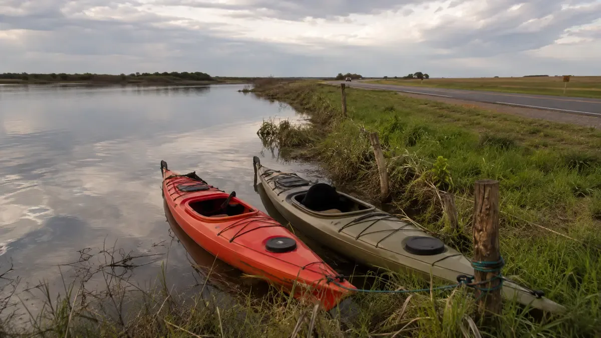 Kayaks genéricos amarrados cerca de la costa en una ruta entrerriana entre campos, plano medio documental, perspectiva natural