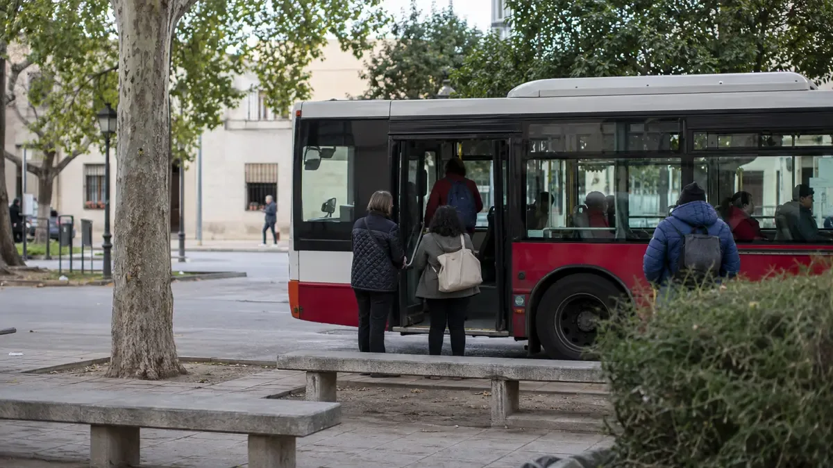 Un colectivo urbano genérico detenido en una parada en una plaza de barrio con mobiliario urbano, plano medio documental, perspectiva natural